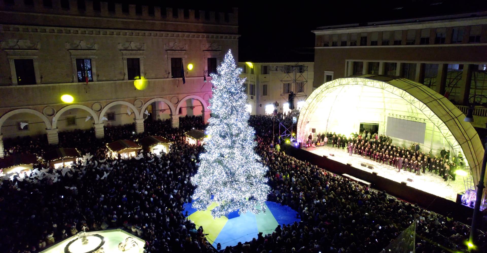 Albero di Natale illuminato con folla e palco in piazza.